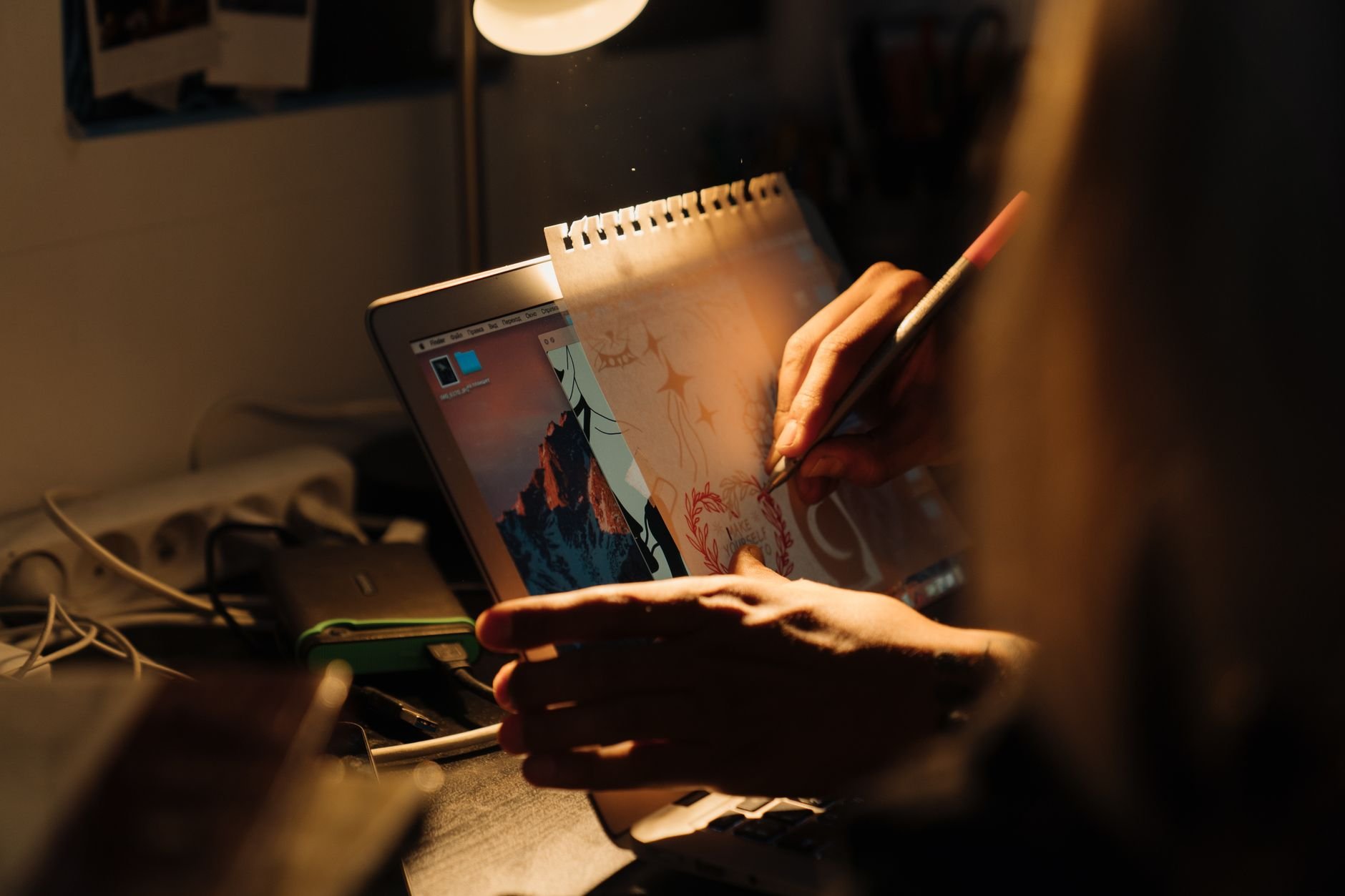 Artist working on a design using a laptop and paper under warm lighting indoors.